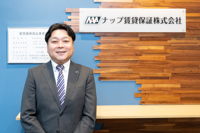 ナップ賃貸保証株式会社のロゴの前でインタビューを受けるナップ賃貸保証株式会社の専務取締役の田邊氏
