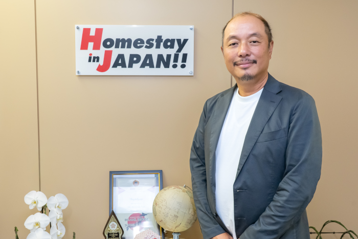 株式会社ネクステージのロゴの前に立つ代表取締役、梶田佳彦氏