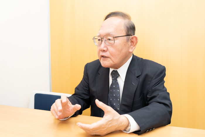 信明ゼネラル株式会社のロゴの前でインタビューを受ける信明ゼネラル株式会社の代表取締役の田中光一氏