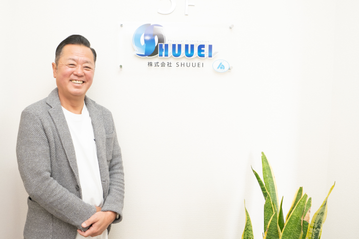 shuueiのロゴの前でインタビューを受ける株式会社shuueiの代表取締役社長菊池氏