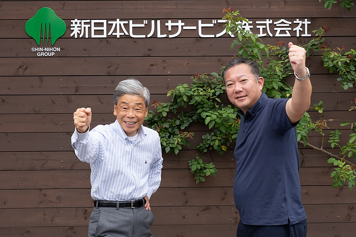 新日本ビルサービス株式会社の社名看板の前でガッツポーズをとる関根一成氏と照井洋輔氏